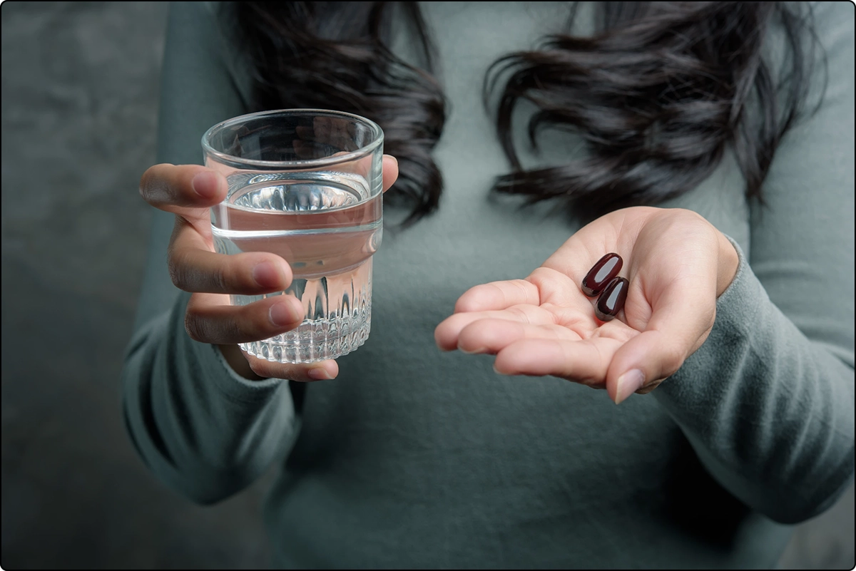 woman with blue grey sweater holding glass of water and two capsules, avoid pill fatigue