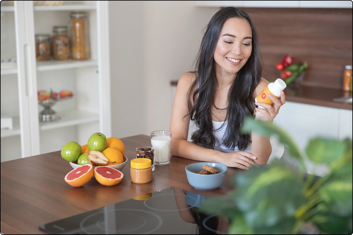attractive woman reading ingredients on multivitamin supplement bottle in her modern kitchen with fruits on the counter