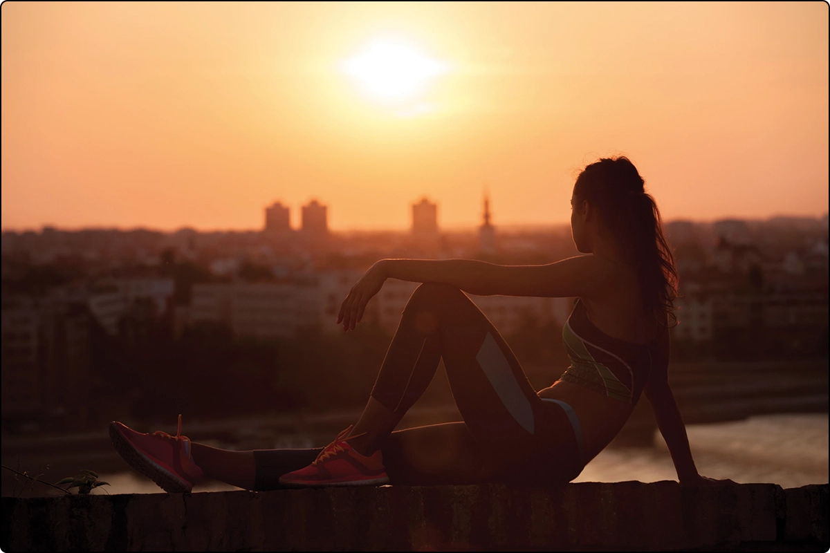 young woman in athletic wear resting on ledge looking past_a town to the glowing orange sunset