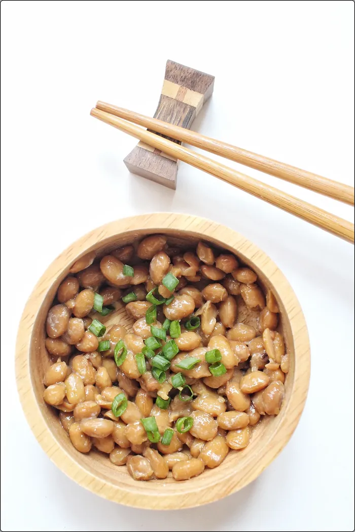 natto topped with chopped chives in a wooden bowl next to wooden chopsticks